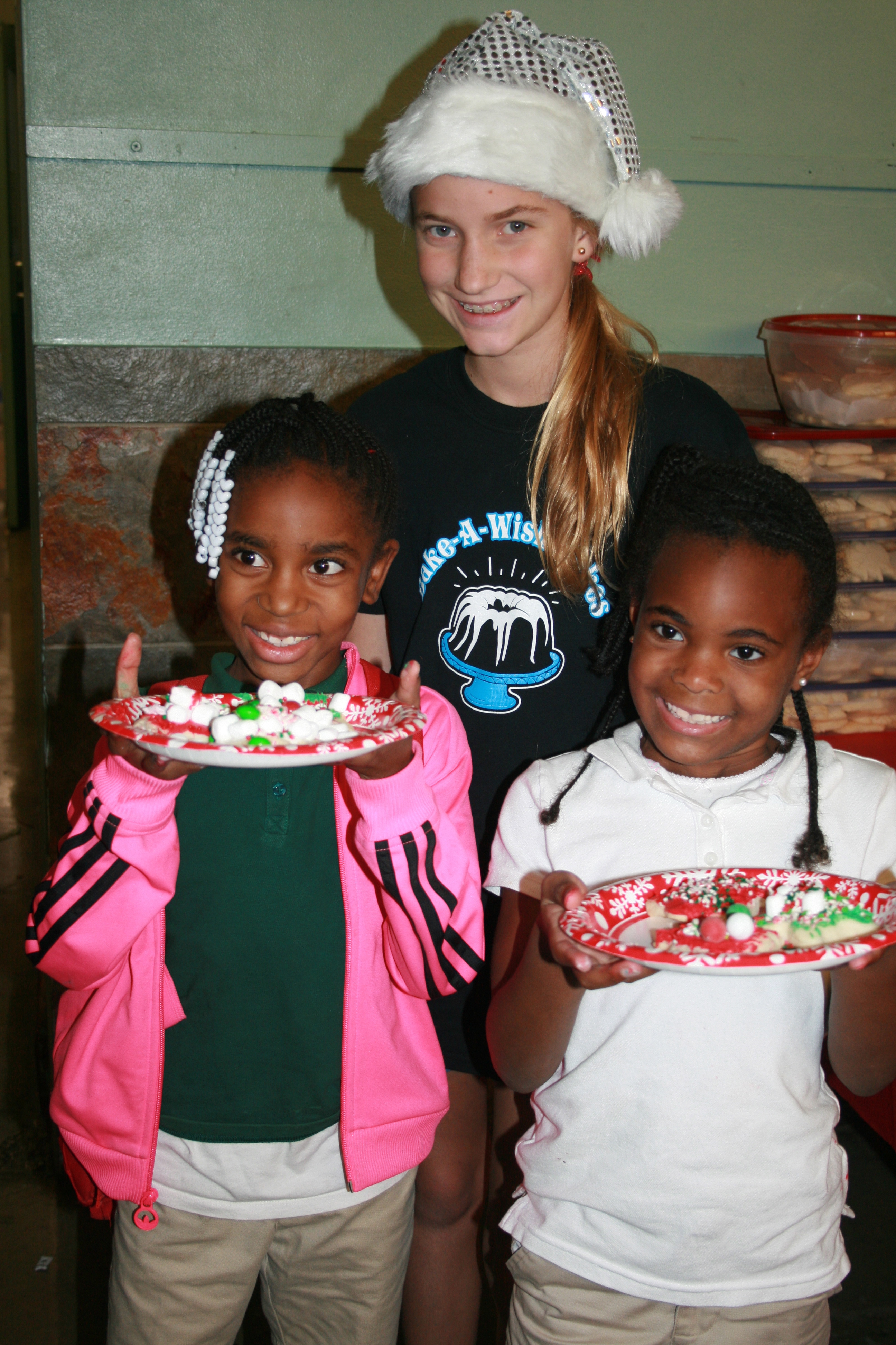 Volunteer in sparkly holiday hat with two children holding decorated cookie plates