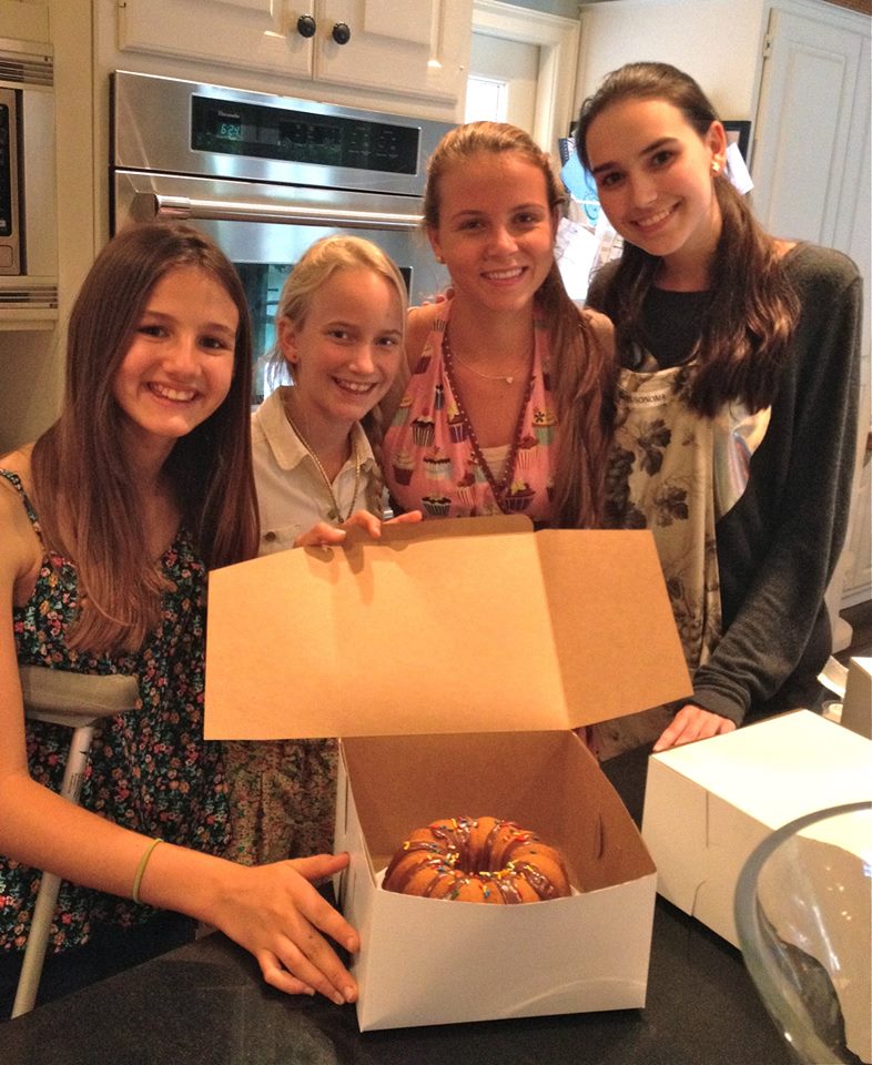Four young female volunteers in kitchen setting holding open cake box with decorated bundt cake