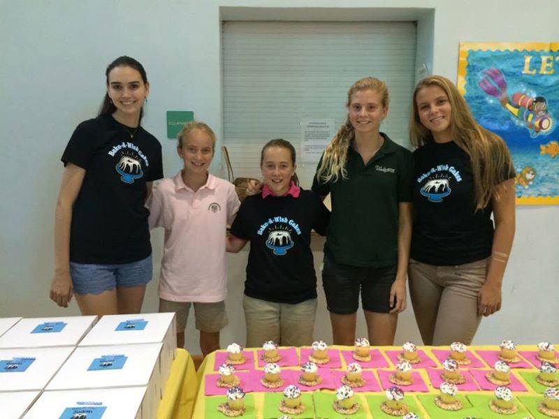 Five volunteers standing behind table with rows of decorated cupcakes on colorful mats