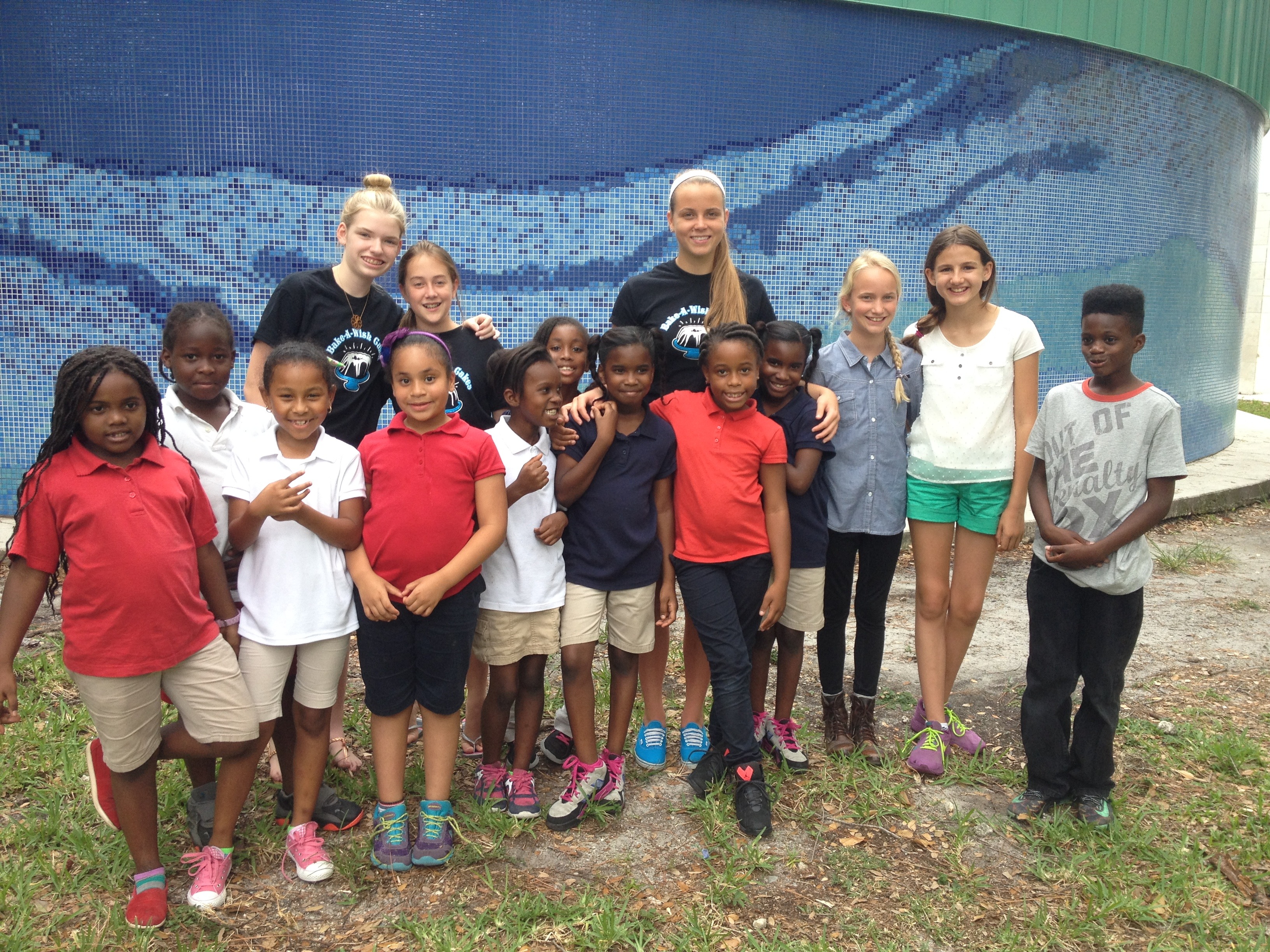Large group photo of Bake-A-Wish volunteers with children in school uniforms outdoors by blue tiled wall